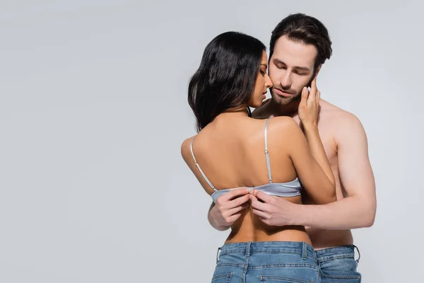 Молодой мужчина без рубашки расстегивает бюстгальтер сексуальной женщины, изолированный на серой — стоковое фото