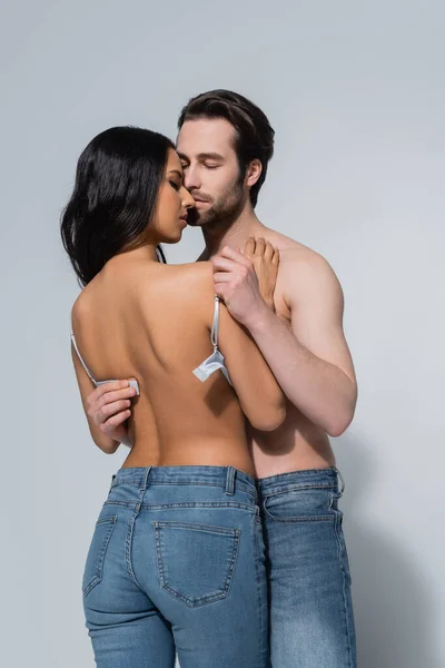 Hemdloser Mann mit geschlossenen Augen zieht sexy Frau in Jeans auf grau den BH aus — Stockfoto