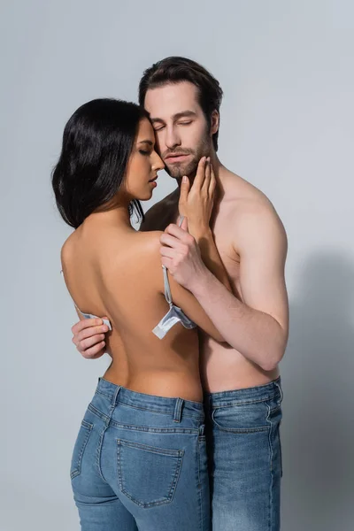 Young shirtless man holding bra of sexy woman touching his face on grey — Stock Photo