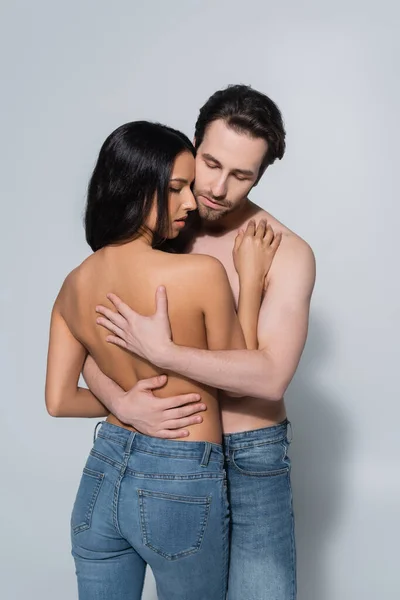Joven hombre sin camisa con los ojos cerrados abrazando a la mujer en jeans en gris - foto de stock