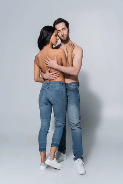 Vista a figura intera di donna e uomo senza camicia in jeans che si abbraccia sul grigio — Foto stock