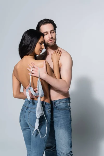 Sexy shirtless man holding bra while embracing seductive brunette woman on grey — Stock Photo
