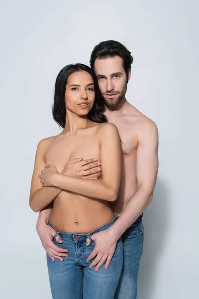Woman covering breast with hands near shirtless man holding thumbs in belt loops of her jeans on grey — Stock Photo