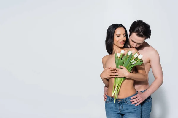 Femme souriante en jeans ne tenant des tulipes près sexy homme torse nu sur gris — Photo de stock