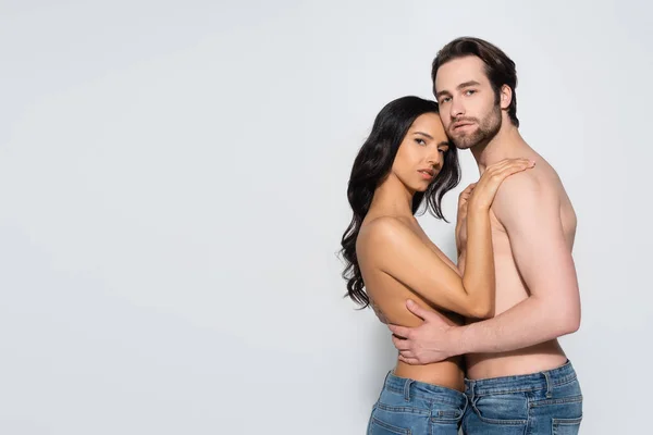 Sexy joven pareja en jeans mirando a la cámara mientras se abraza en gris — Stock Photo