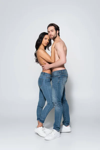 Full length view of young couple in jeans and sneakers looking at camera while embracing on grey — Stock Photo