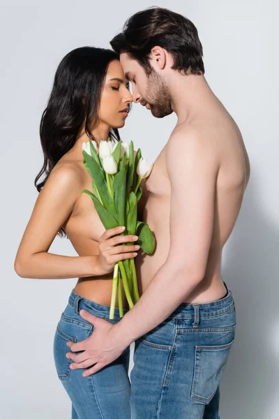 Сексуальная брюнетка с тюльпанами и мужчиной без рубашки в джинсах, стоящим лицом к лицу с закрытыми глазами на сером — стоковое фото