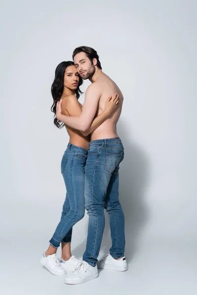 Vista completa de pareja sexy en jeans y zapatillas de deporte mirando a la cámara mientras se abraza en gris - foto de stock