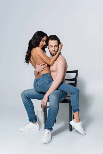 Homme torse nu en jeans assis sur une chaise avec une petite amie passionnée sur gris — Photo de stock