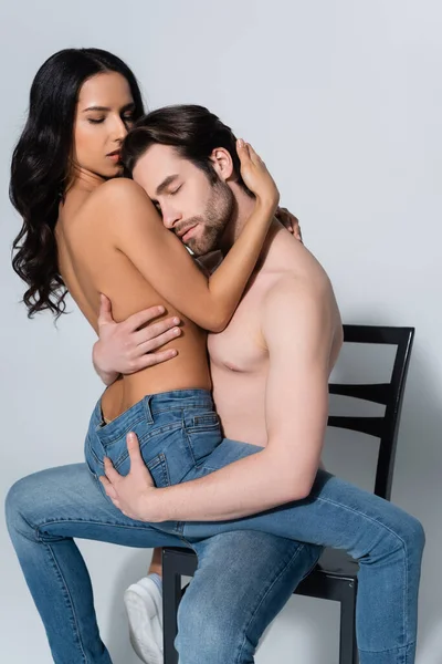 Shirtless man with closed eyes hugging woman in jeans while sitting on chair on grey — Stock Photo