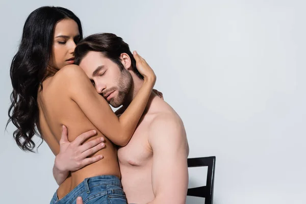 Hombre sin camisa con los ojos cerrados sentado en la silla y abrazando a la mujer morena aislado en gris - foto de stock