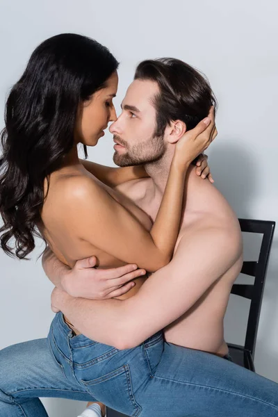 Homme torse nu embrassant femme brune passionnée tout en étant assis sur une chaise sur gris — Photo de stock