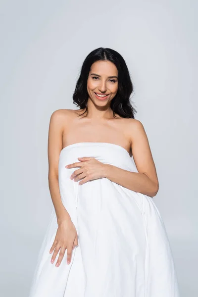 Brunette woman in white blanket looking at camera isolated on grey — Stock Photo