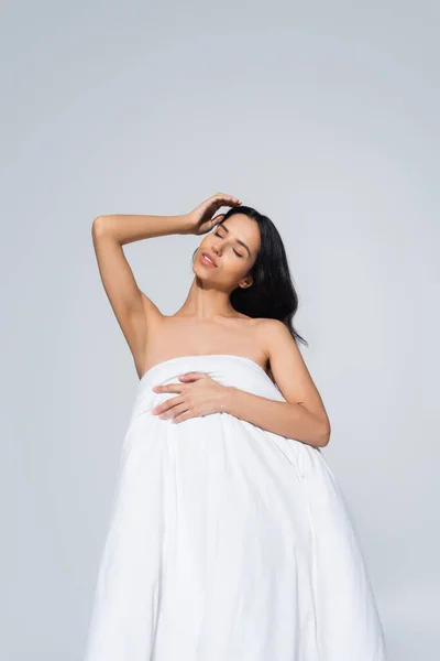 Young woman with naked shoulders and closed eyes posing with white blanket isolated on grey — Stock Photo