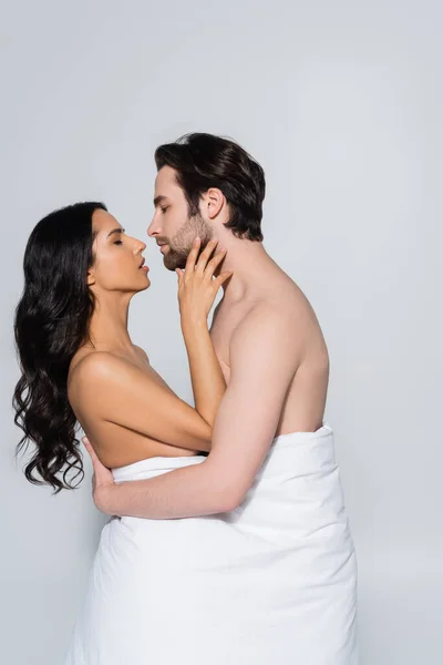 Vista lateral del hombre sin camisa y la mujer apasionada en manta blanca abrazando mientras que de pie cara a cara aislado en gris - foto de stock