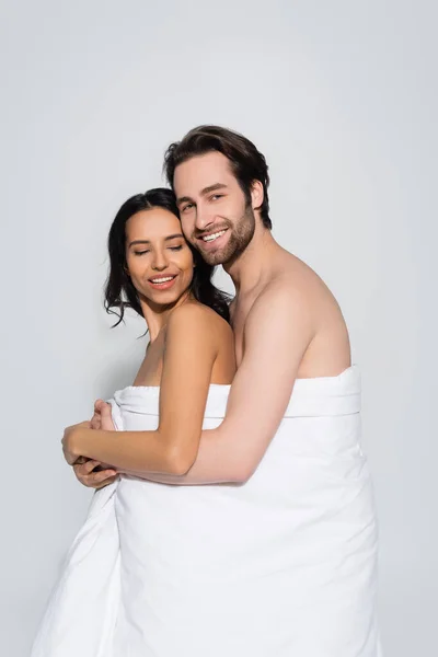 Cheerful man looking at camera while hugging sexy woman in white blanket  isolated on grey — Stock Photo