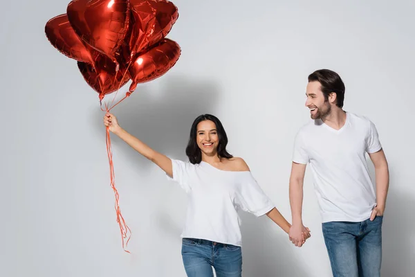 Cheerful woman with heart-shaped balloons holding hands with young boyfriend on grey — Stock Photo