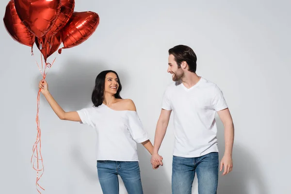 Heureux jeune couple en t-shirts blancs tenant la main et se regardant près de ballons en forme de coeur sur gris — Photo de stock