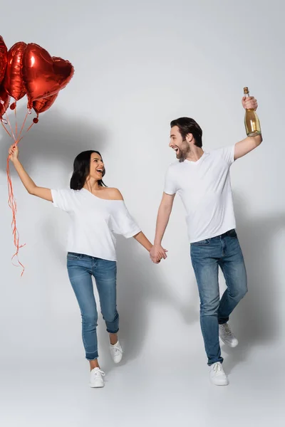 Ganzkörperansicht des glücklichen Paares beim Gehen mit roten herzförmigen Luftballons auf grau — Stockfoto