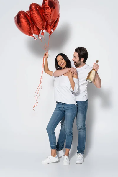 Felice uomo con bottiglia di champagne abbracciando fidanzata con palloncini a forma di cuore su grigio — Foto stock