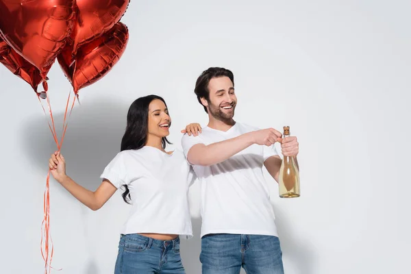 Homme souriant ouvrant bouteille de champagne près de femme heureuse avec des ballons rouges sur gris — Photo de stock