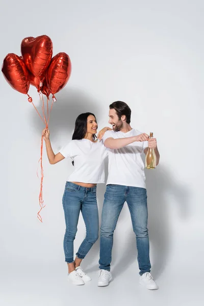 Allegro uomo apertura bottiglia di champagne vicino fidanzata con palloncini a forma di cuore su grigio — Foto stock