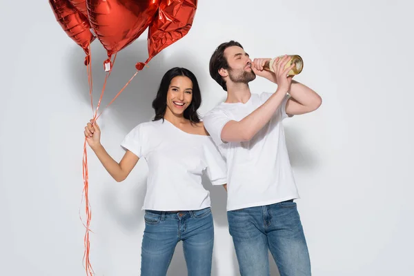 Cheerful woman with red balloons looking at camera near man drinking champagne from bottle on grey — Stock Photo