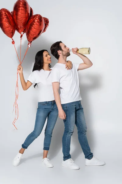 Piena vista della donna gioiosa con palloncini rossi a forma di cuore vicino all'uomo che beve champagne dalla bottiglia sul grigio — Foto stock