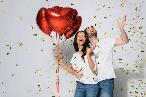 Cheerful couple with heart-shaped balloons laughing under falling confetti on grey — Stock Photo