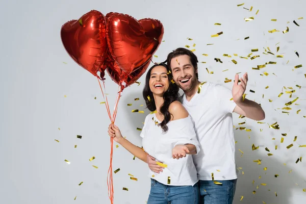 Happy woman with red heart-shaped balloons smiling near boyfriend and confetti on grey — Stock Photo