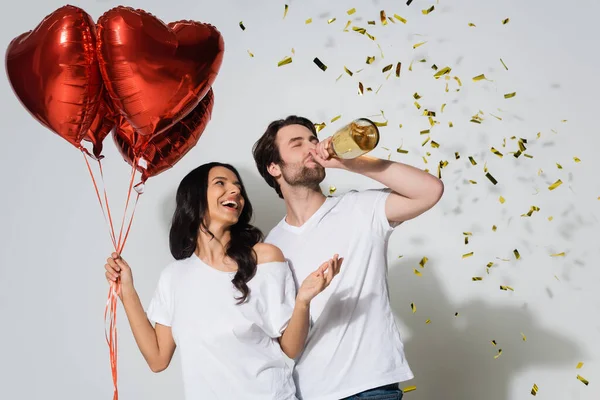 Mujer excitada sosteniendo globos en forma de corazón y el hombre bebiendo champán de la botella cerca de confeti en gris - foto de stock