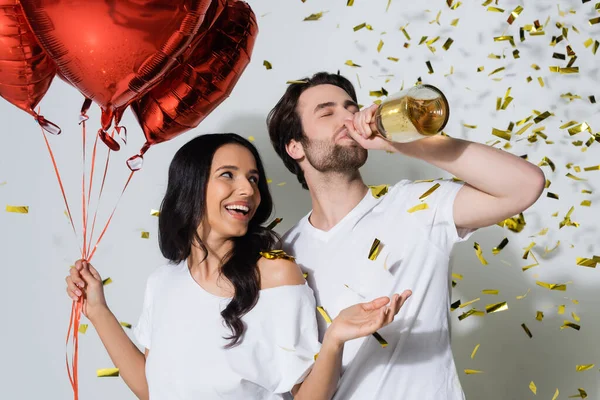 Mujer alegre sosteniendo globos rojos cerca de novio bebiendo champán de botella en gris - foto de stock