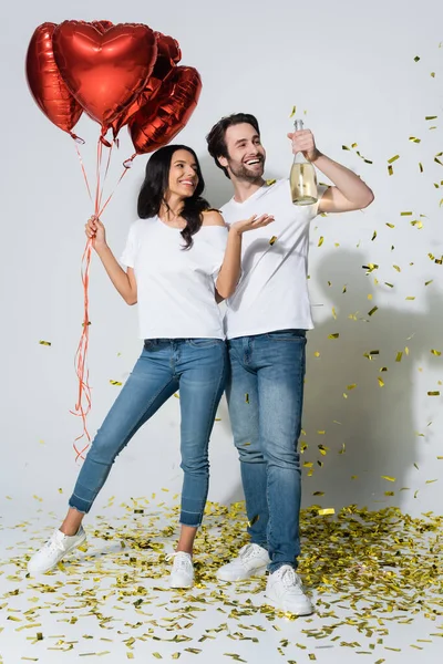 Mujer feliz con globos en forma de corazón apuntando a la botella de champán en la mano del novio en gris - foto de stock