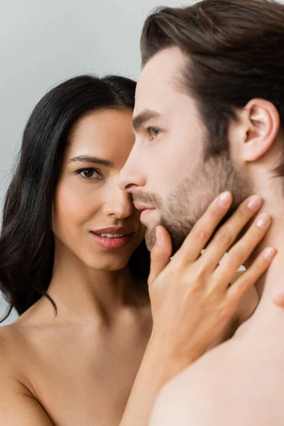 Young passionate woman looking at camera while touching face of young man isolated on grey — Stock Photo