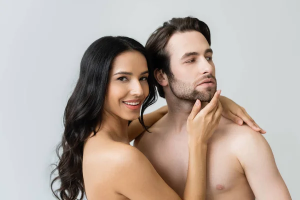 Улыбающаяся брюнетка женщина трогает лицо молодого человека, изолированного на сером — стоковое фото