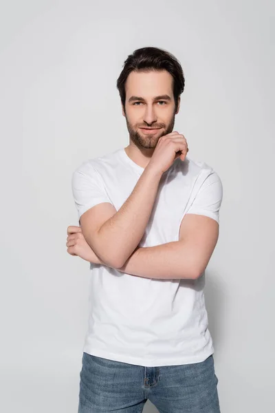 Positive man in white t-shirt holding hand near face while looking at camera on grey — Stock Photo