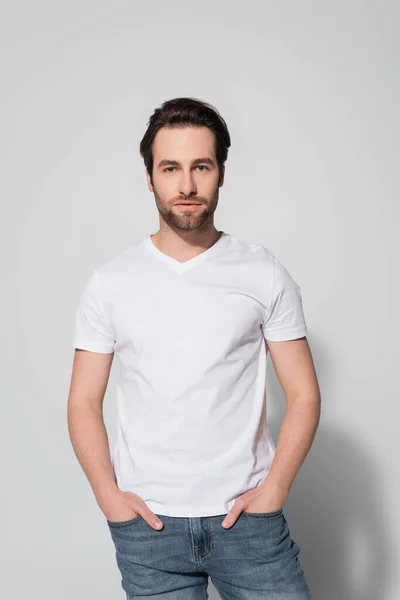 Brunette man in white t-shirt standing with hands in pockets of jeans on grey — Stock Photo