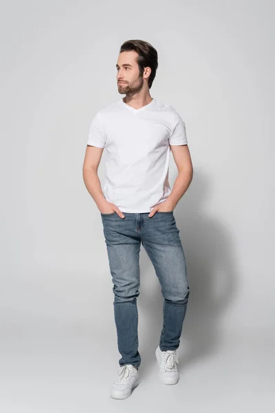 Full length view of young man in white t-shirt looking away while standing with hands in pockets of jeans on grey — Stock Photo