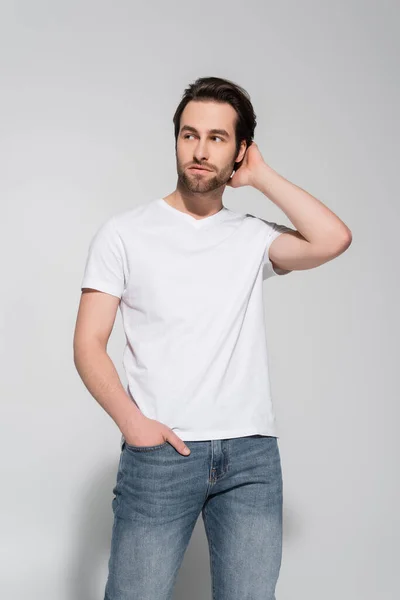 Young man in white t-shirt holding hand in pocket of jeans, touching hair and looking away on grey — Stock Photo