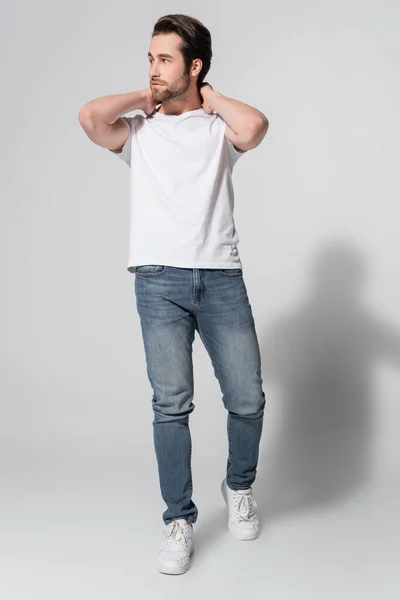 Full length view of young man in jeans and white t-shirt posing with hands behind neck on grey — Stock Photo