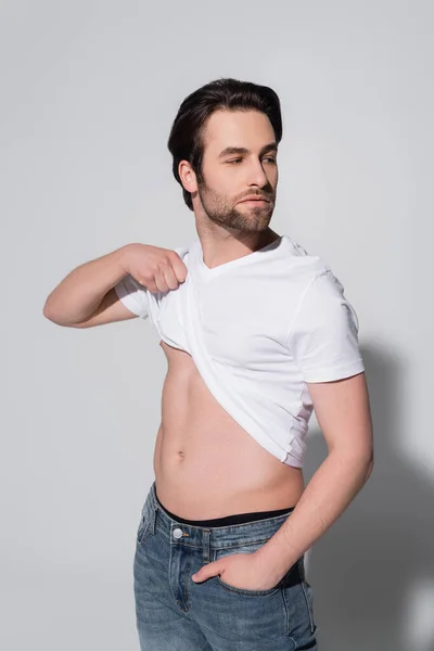 Sexy man raising white t-shirt while standing with hand in pocket of jeans on grey — стоковое фото
