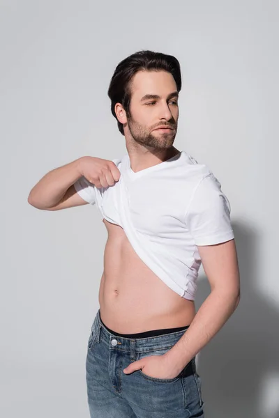 Sexy man raising white t-shirt while standing with hand in pocket of jeans on grey — Stock Photo