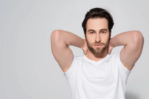 Brünetten bärtigen Mann in weißem T-Shirt hält die Hände hinter dem Hals, während Blick in die Kamera auf grau — Stockfoto