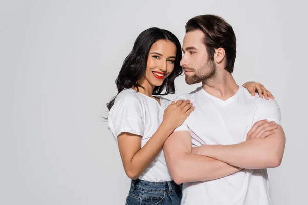 Sorridente donna bruna che guarda la macchina fotografica mentre abbraccia le spalle dell'uomo in piedi con le braccia incrociate isolate sul grigio — Foto stock