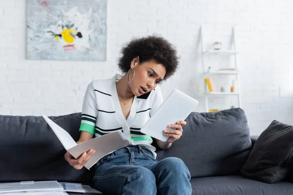 Mujer afroamericana enfocada mirando la tableta digital, hablando en el teléfono inteligente y sosteniendo la carpeta de papel en casa - foto de stock