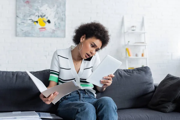 Focalizzato donna afroamericana guardando tablet digitale, parlando su smartphone e tenendo cartella di carta a casa — Foto stock