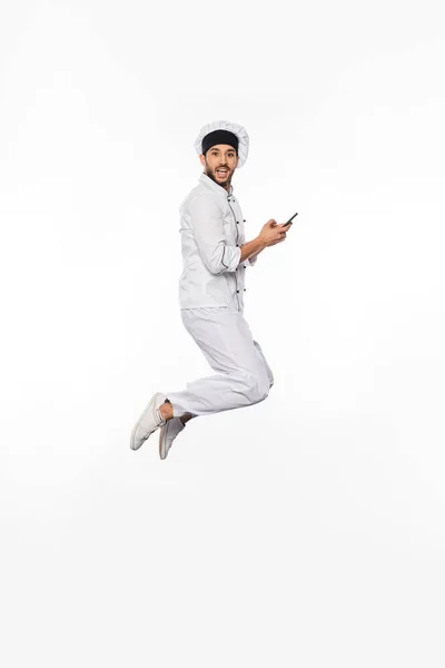 Здивований шеф-кухар в капелюсі і рівномірний стрибок зі смартфоном на білому — стокове фото