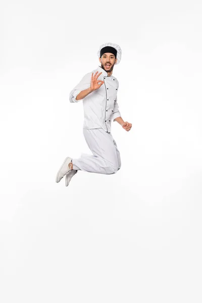 Asombrado chef en sombrero y uniforme saltando y mostrando bien en blanco - foto de stock