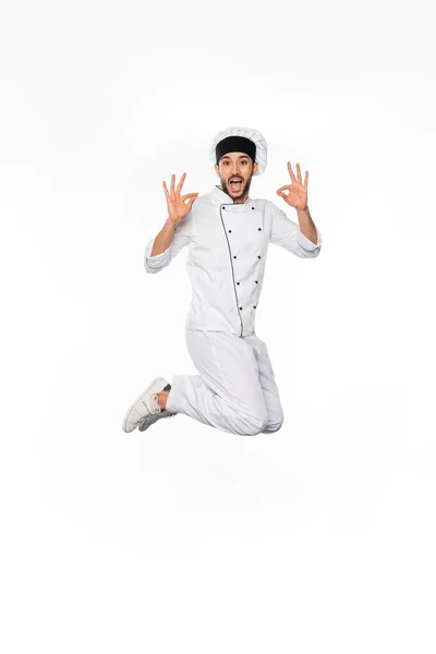 Chef emocionado en sombrero y uniforme saltando y mostrando bien en blanco - foto de stock