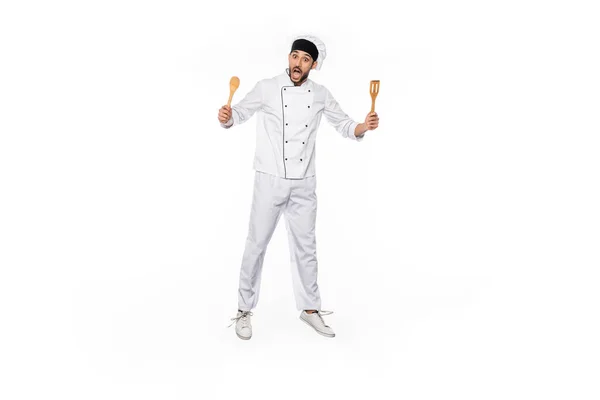 Chef sorprendido en sombrero y uniforme saltando y sosteniendo espátula de madera y cuchara aislada en blanco - foto de stock